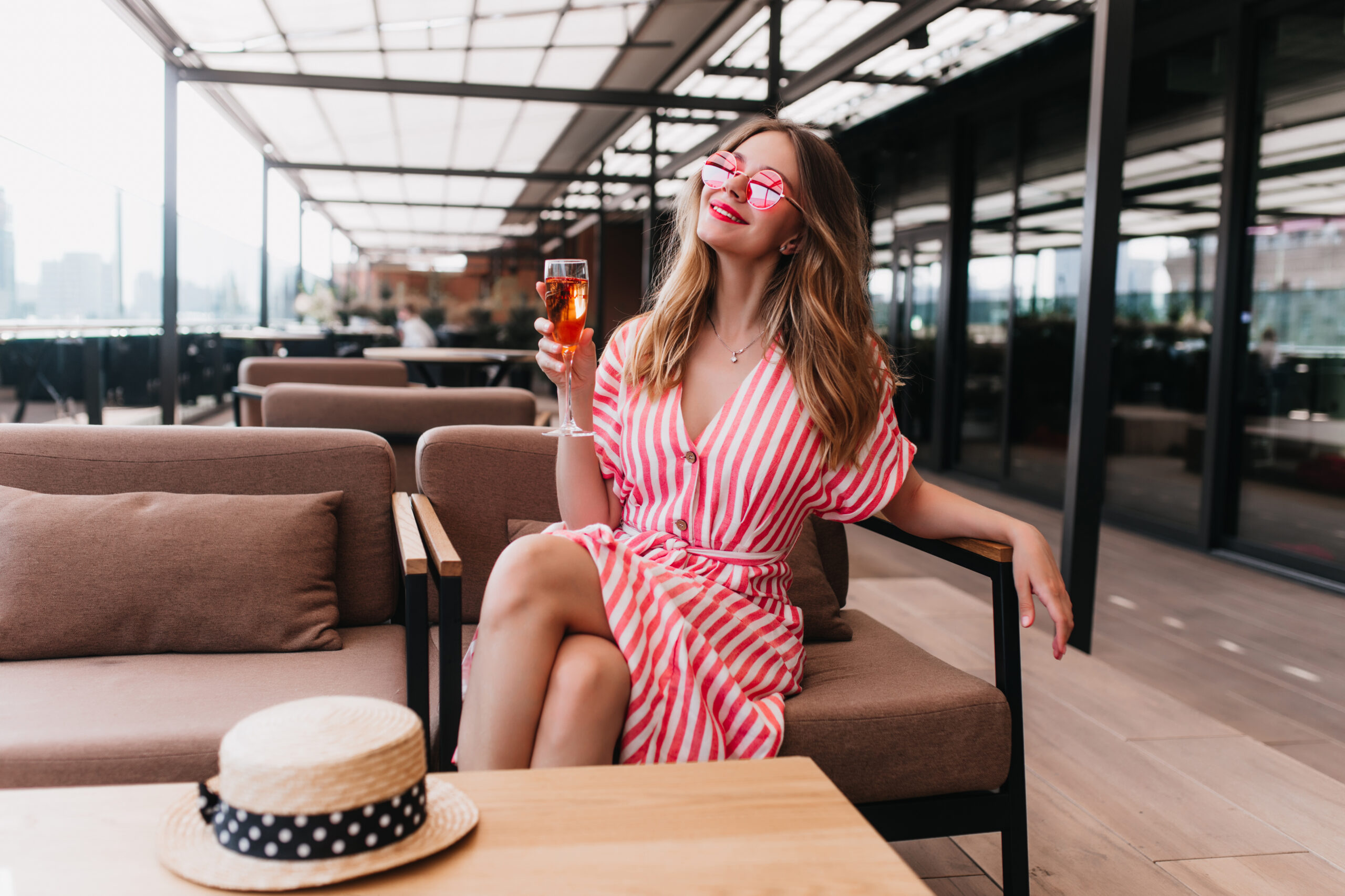 Woman wearing a pink and white striped summer dress with sunglasses, holding a cocktail on a cruise deck lounge area.